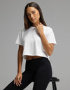 A woman models the Cozy Earth Women’s All Day Cropped Tee in white and black leggings, seated on a stool and looking at the camera with a neutral expression against a plain gray background.|Color:White