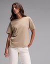 A person stands against a plain gray background, wearing the Cozy Earth Women’s All Day Tee in Beech Heather paired with white pants. They have long brown hair and a relaxed pose, looking slightly to the side.|Color:Beech Heather