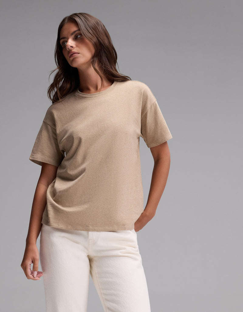 A person stands against a plain gray background, wearing the Cozy Earth Women’s All Day Tee in Beech Heather paired with white pants. They have long brown hair and a relaxed pose, looking slightly to the side.