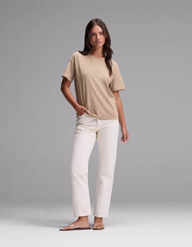 A woman stands against a plain gray background, wearing the Cozy Earth Women’s All Day Tee in beige, paired with white jeans and beige sandals. One hand is in her pocket as she looks slightly to the side.