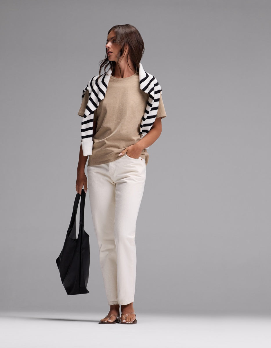 A woman stands against a plain gray background wearing Cozy Earth’s Women’s All Day Tee in beige, paired with white pants, sandals, and a black-and-white striped sweater draped over her shoulders, while holding a large black tote bag.