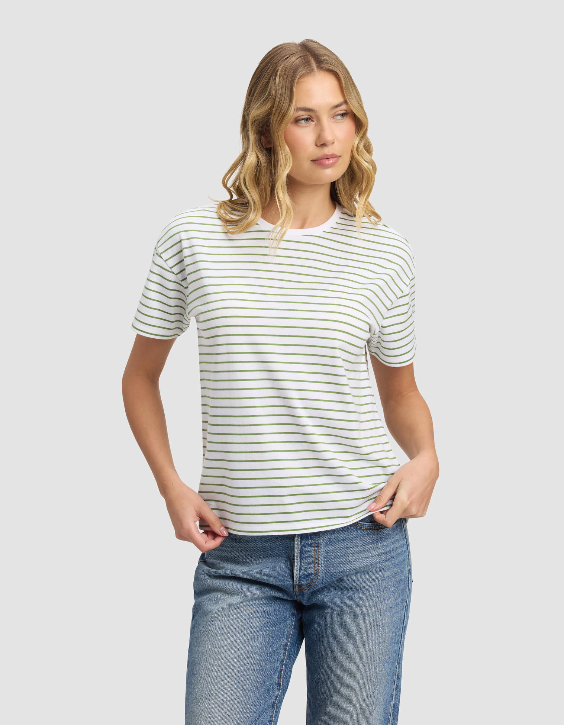 A woman with wavy blonde hair models the Cozy Earth Women's All Day Tee, a white short-sleeve shirt with thin black stripes, paired with blue jeans. She stands against a plain light gray background, gazing to the side in a relaxed mood.|Color:Clover Stripe