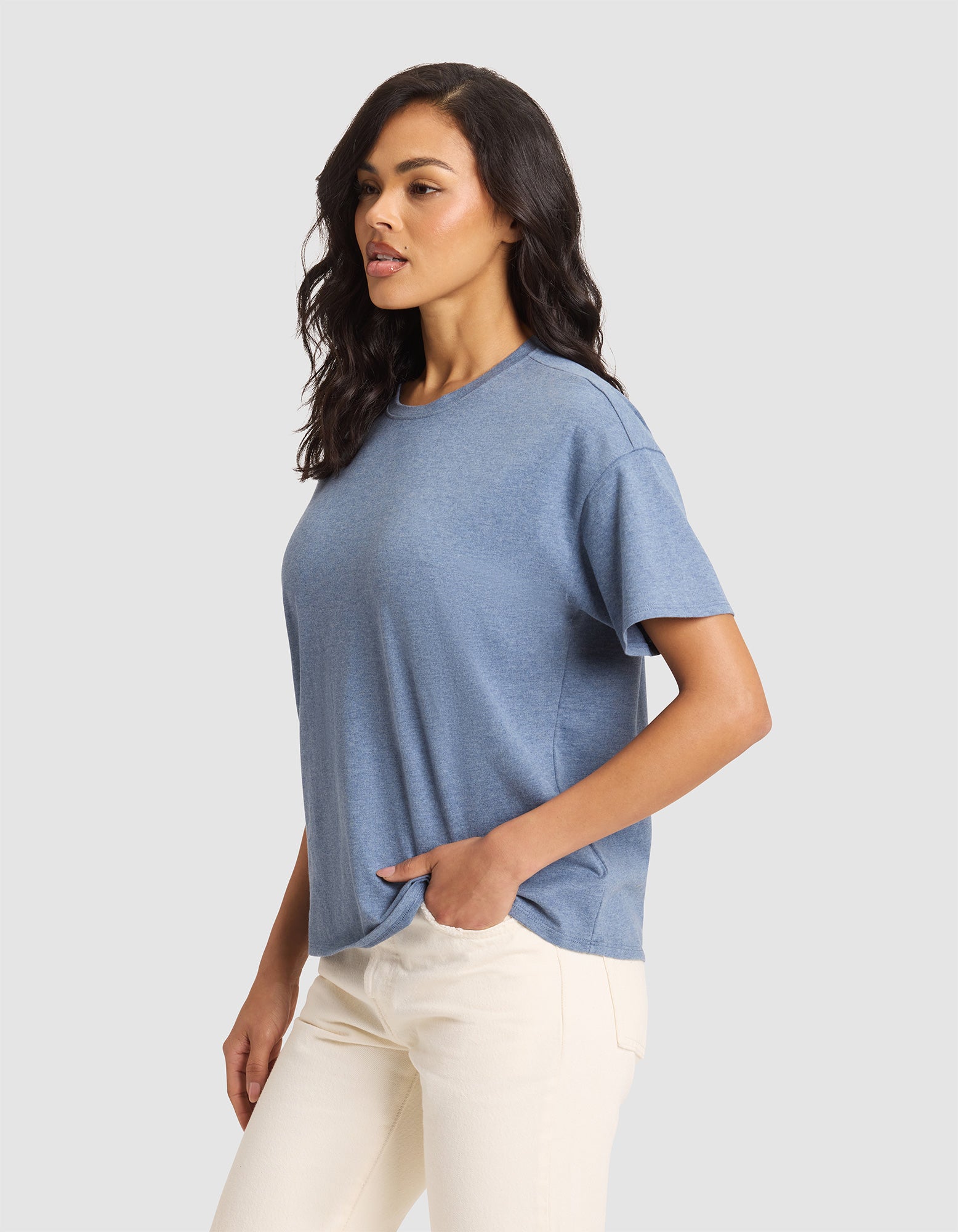 A woman with wavy dark hair models the Cozy Earth Women's All Day Tee, a loose-fitting blue short-sleeve t-shirt, paired with light-colored pants, standing with one hand in her pocket against a plain light gray background.|Color:Deep Blue Heather