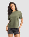 A woman with long dark hair wears the Cozy Earth Women's All Day Tee in green and black denim shorts, standing against a plain light gray background facing forward with one hand on her hip. |Color:Olive Heather