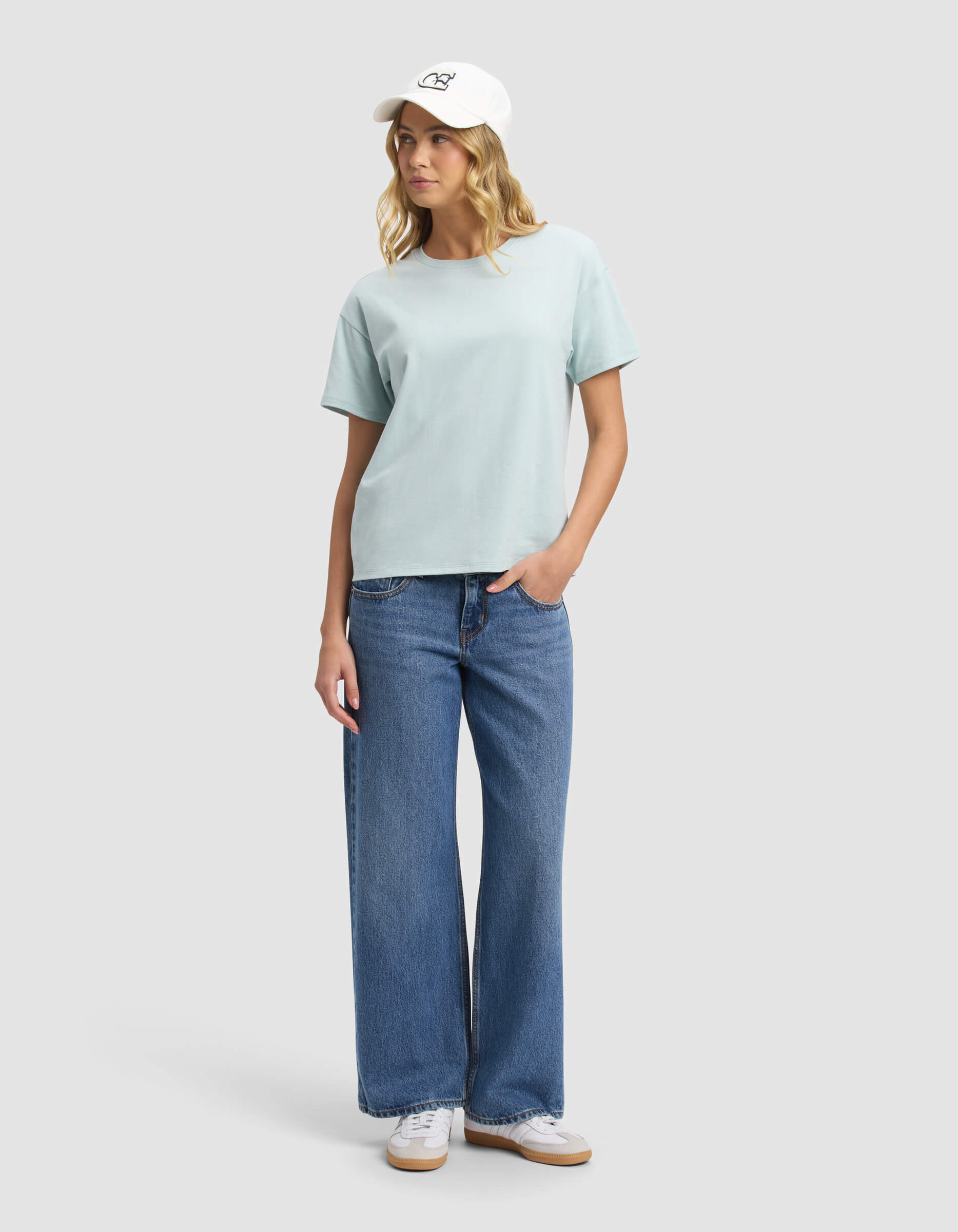 A person stands against a plain light gray background, wearing Cozy Earth’s Women's All Day Tee in light blue, blue wide-leg jeans, white sneakers, and a white baseball cap, looking slightly to the side with one hand in their pocket.