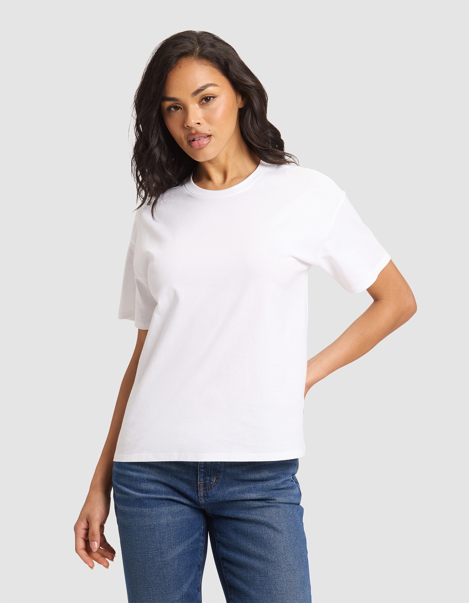 A woman models the Cozy Earth Women's All Day Tee in plain white, paired with blue jeans, standing against a light gray background with one hand on her hip and a neutral expression.  