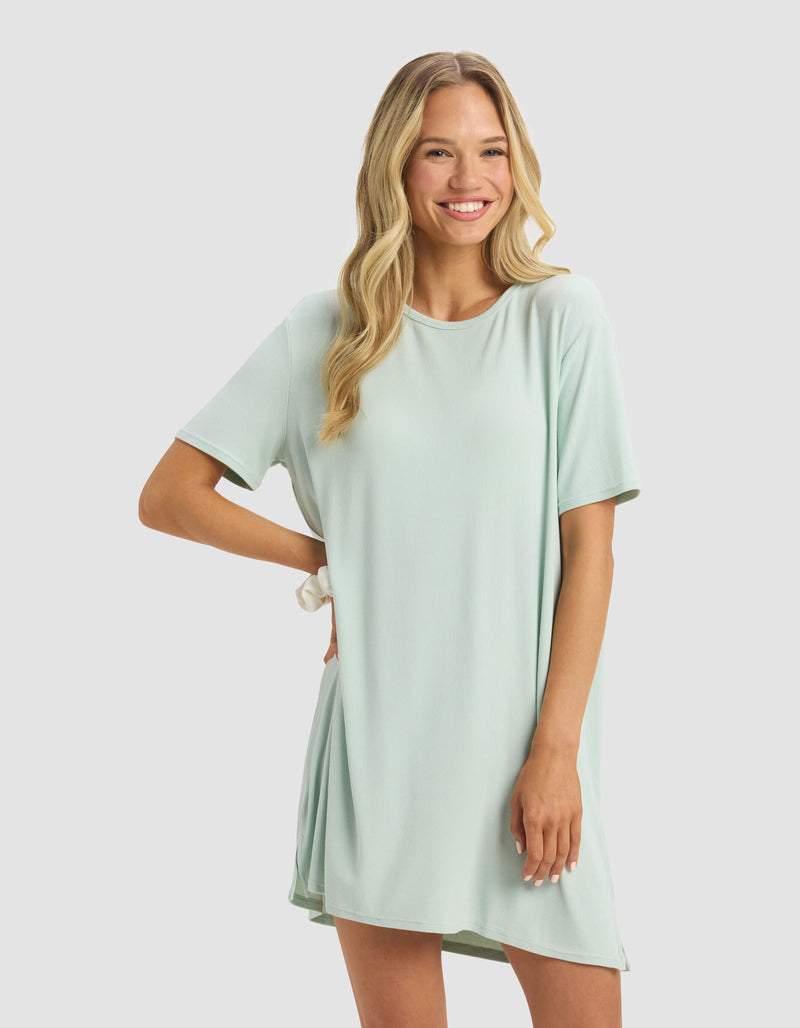 A smiling woman with long blonde hair poses with one hand on her hip against a light gray background, wearing the Cozy Earth Women's Bamboo Rib-Knit Boyfriend Dress in light green.