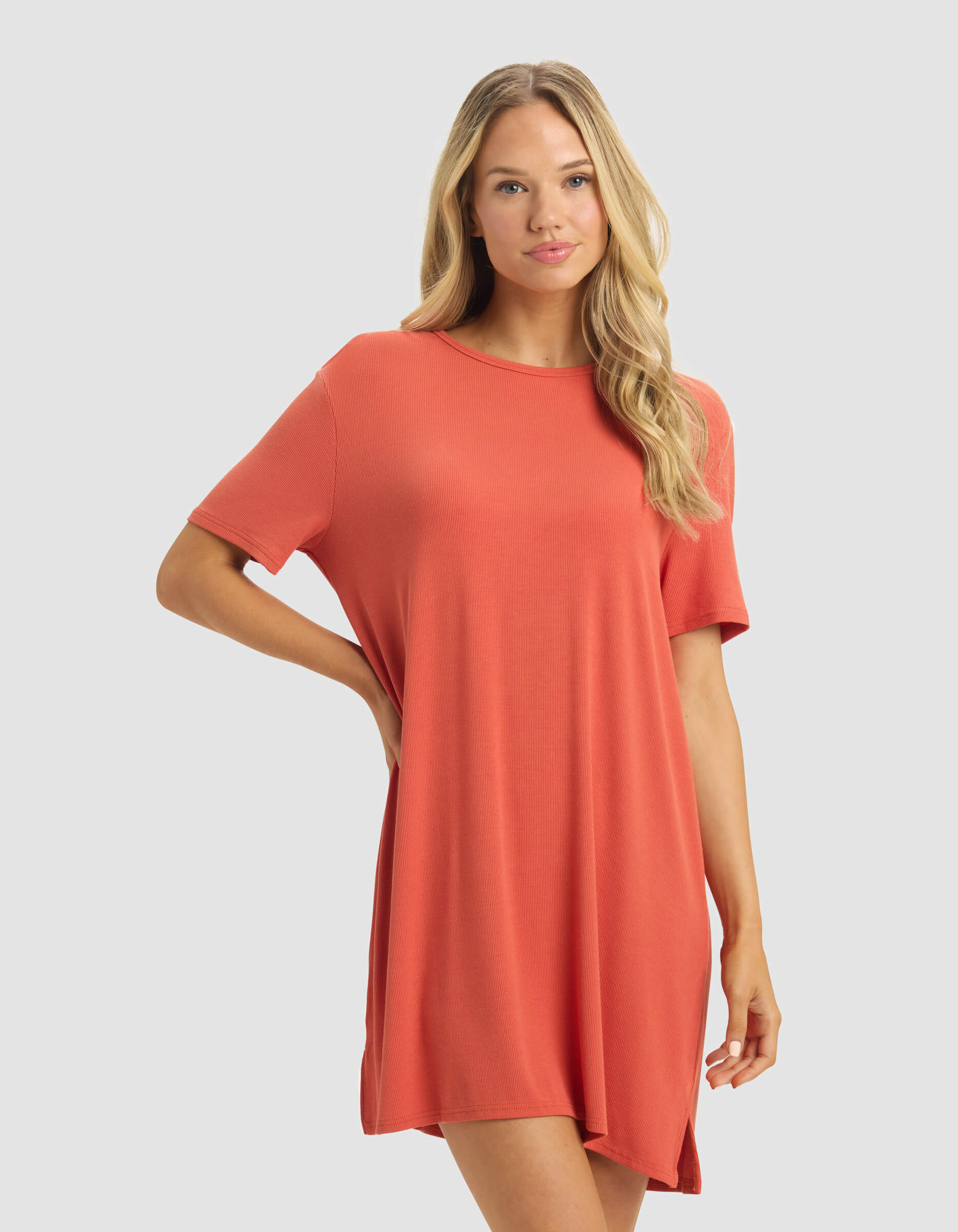 A woman with long blonde hair wears the Cozy Earth Women's Bamboo Rib-Knit Boyfriend Dress in coral-orange, standing against a light gray background with one hand on her hip and a neutral expression.
