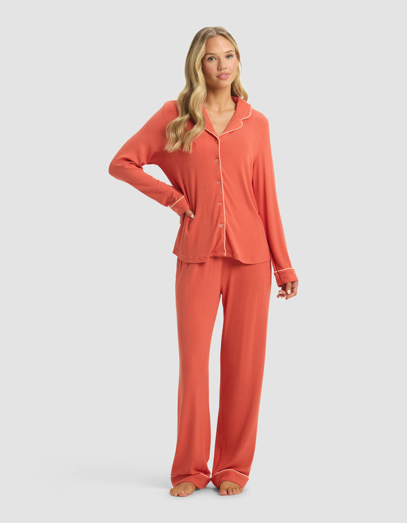 A woman wears the Cozy Earth Women's Bamboo Rib-Knit Classic Long Sleeve Pajama Set, featuring a matching button-up top and long pants in coral. Barefoot, she poses with one hand on her hip against a plain light background.
