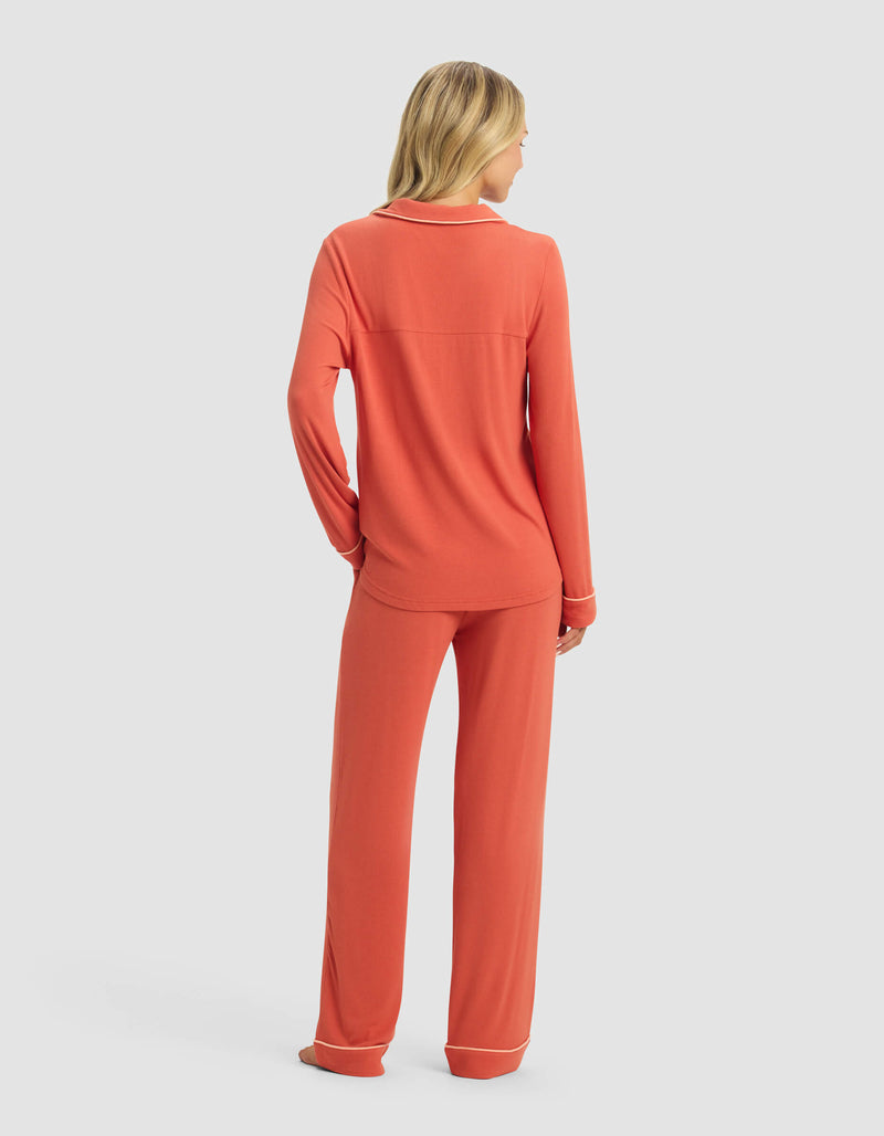 A blonde woman, seen from behind, wears the Cozy Earth Women's Bamboo Rib-Knit Classic Long Sleeve Pajama Set in coral with white trim, standing against a plain light gray background.