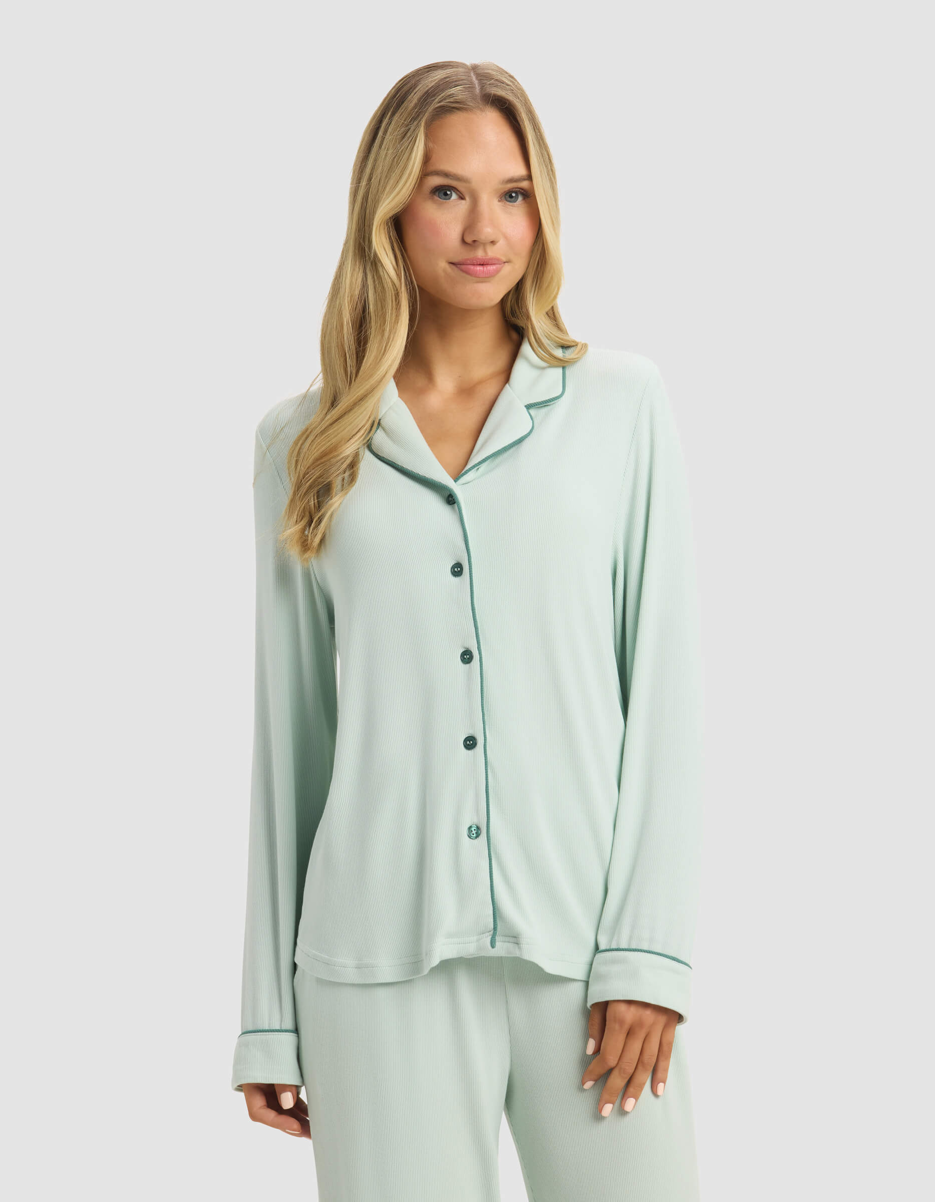 A woman with long blonde hair models Cozy Earth's Women's Bamboo Rib-Knit Classic Long Sleeve Pajama Top in light green with dark green piping, paired with matching pants, against a plain light background.