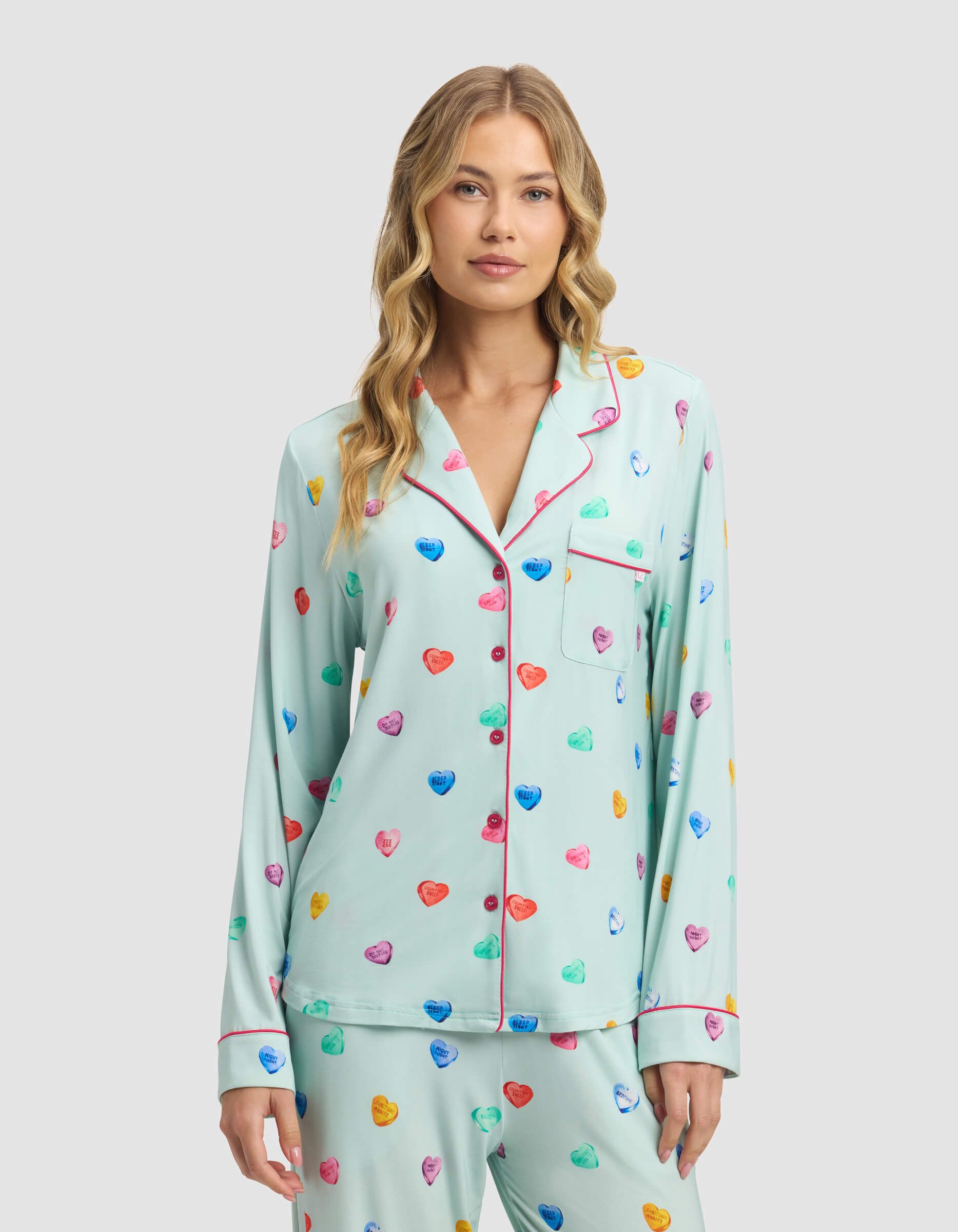 A woman with wavy blonde hair wears the Women's Bamboo Stretch-Knit Long Sleeve Pajama Top by Cozy Earth in light blue with colorful heart patterns and pink trim, standing against a plain light gray background.