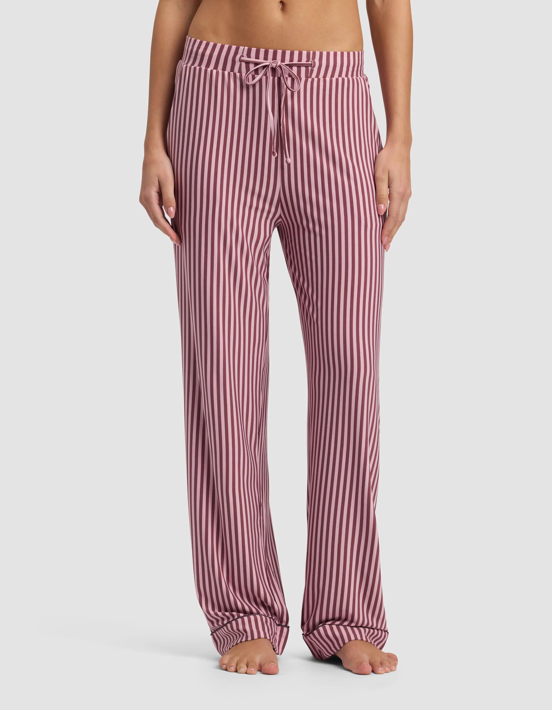 A person stands barefoot, showing only the lower torso and legs, wearing Cozy Earth Women's Bamboo Stretch-Knit Pajama Pant in pink and white vertical stripes with a drawstring waist against a plain background.|Color:Market Stripe in Mulberry