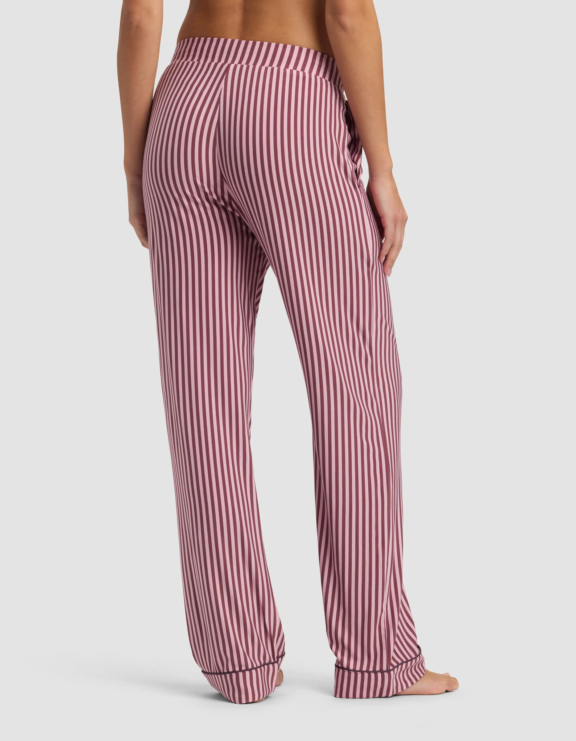 A woman stands barefoot on a white background, shown from the back, wearing Cozy Earth's Women's Bamboo Stretch-Knit Pajama Pant with pink-and-white vertical stripes, an elastic waistband, and a relaxed fit.|Color:Market Stripe in Mulberry