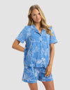 A woman with long blonde hair wears the Cozy Earth Women's Bamboo Stretch-Knit Short Sleeve Pajama Top in blue with white floral patterns, paired with matching shorts, standing and smiling against a plain light gray background.|Color:Garden Toile in Sailor