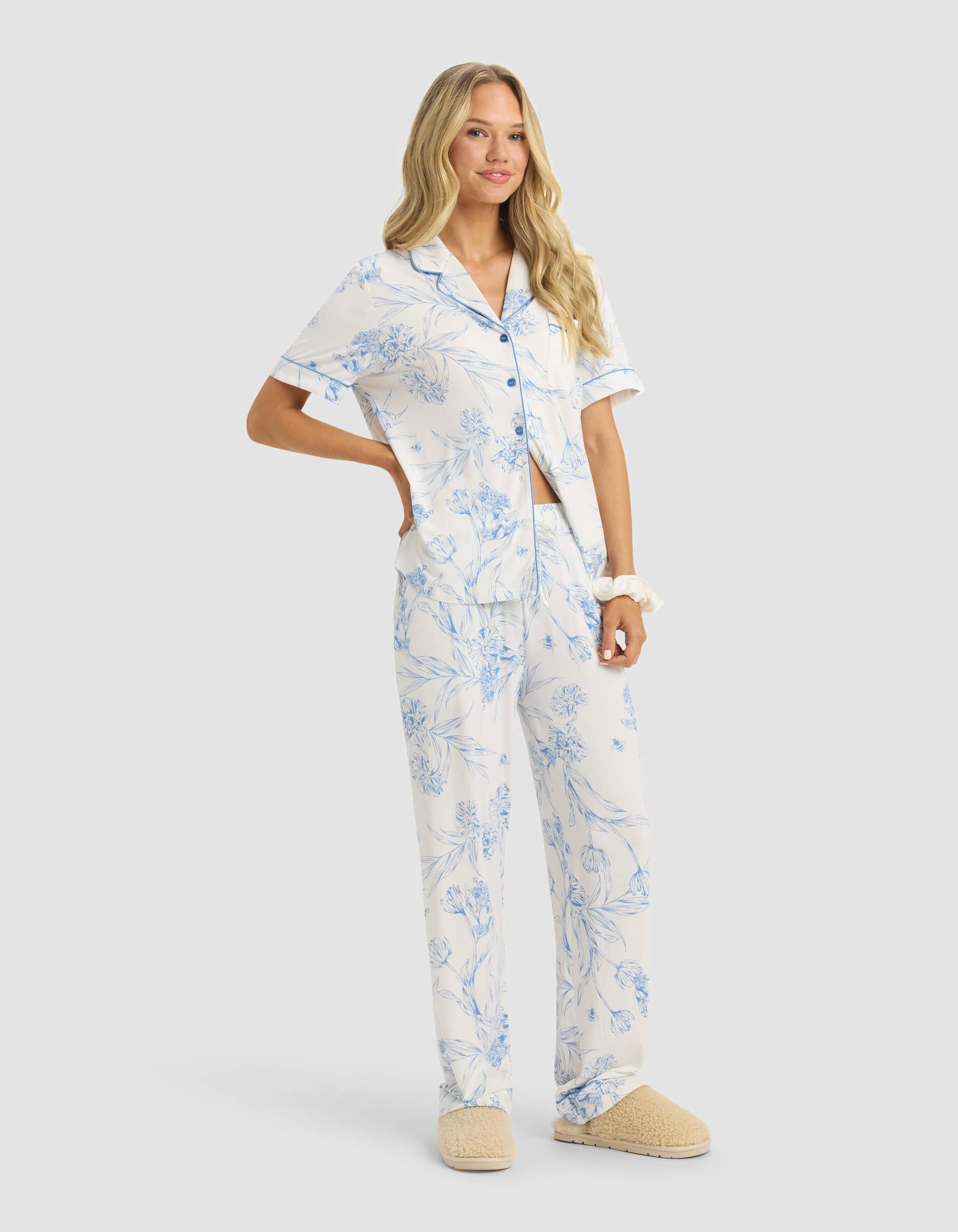 A woman with long blonde hair wears the Cozy Earth Women's Bamboo Stretch-Knit Short Sleeve & Pant Pajama Set in white with blue floral prints and beige slippers, standing casually with one hand on her hip.|Color:Garden Toile in Sea Salt