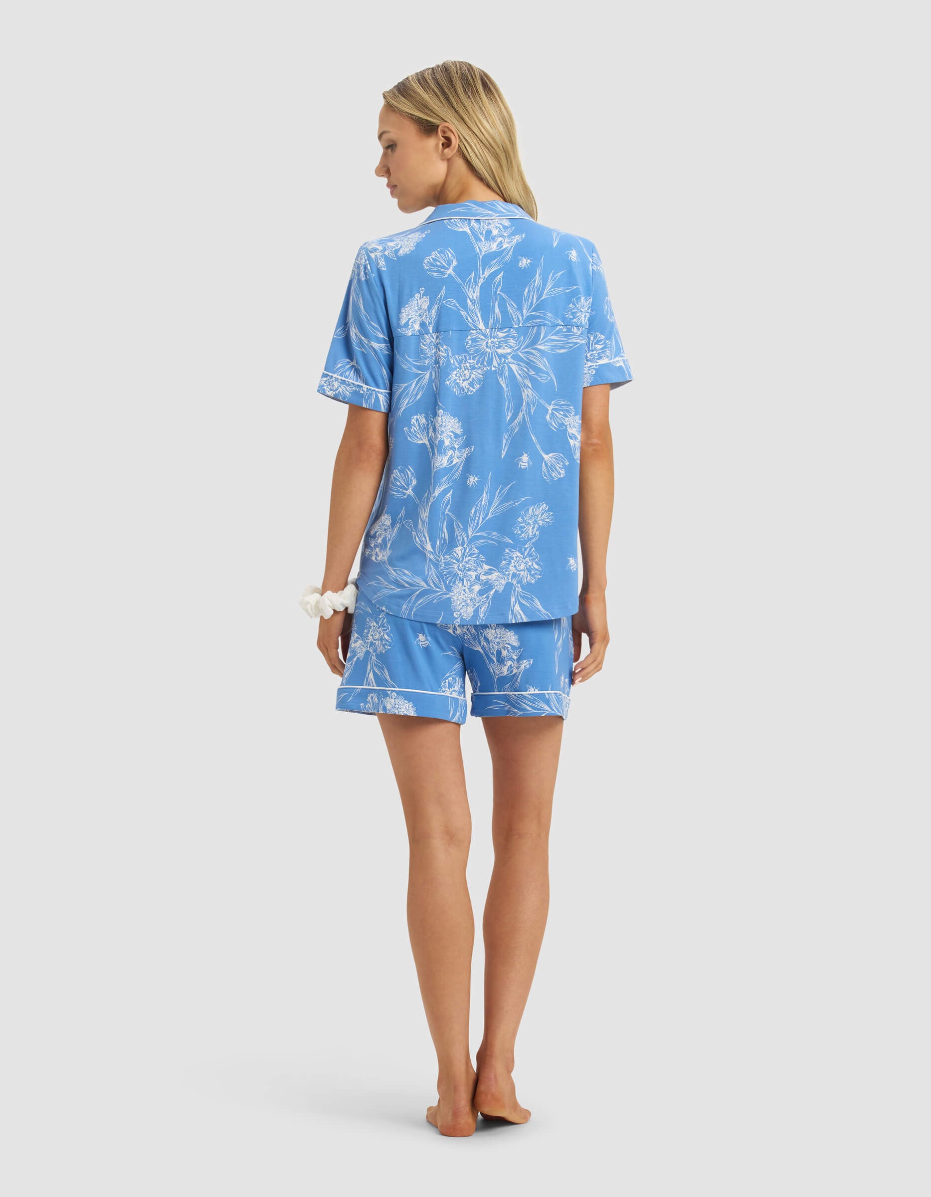 A woman with long blonde hair wears the Cozy Earth Women's Bamboo Stretch-Knit Short Sleeve Pajama Set in light blue with white floral patterns. She stands barefoot facing away, a white scrunchie on her wrist, against a plain white background.