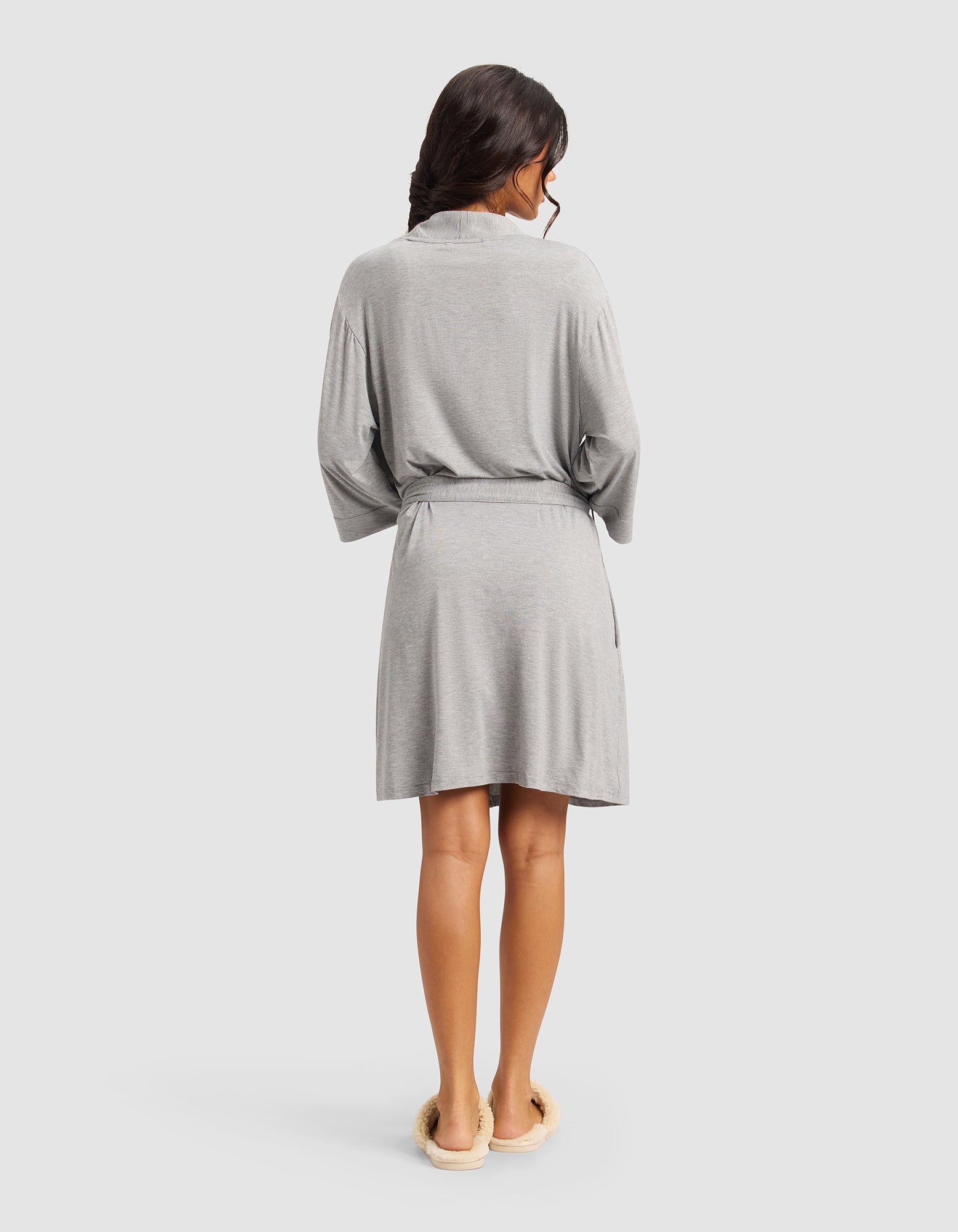 A woman with long dark hair, seen from the back, wears a Cozy Earth Women's Bamboo Stretch-Knit Kimono Robe in light gray with a belt and cream slippers, standing against a plain light background.