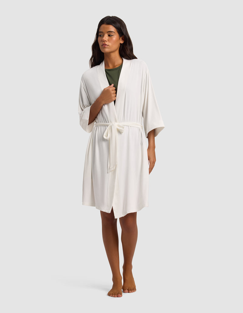 A person stands barefoot against a plain light gray background, wearing Cozy Earth's Women's Bamboo Stretch-Knit Kimono Robe in white with a tie belt over a green top.