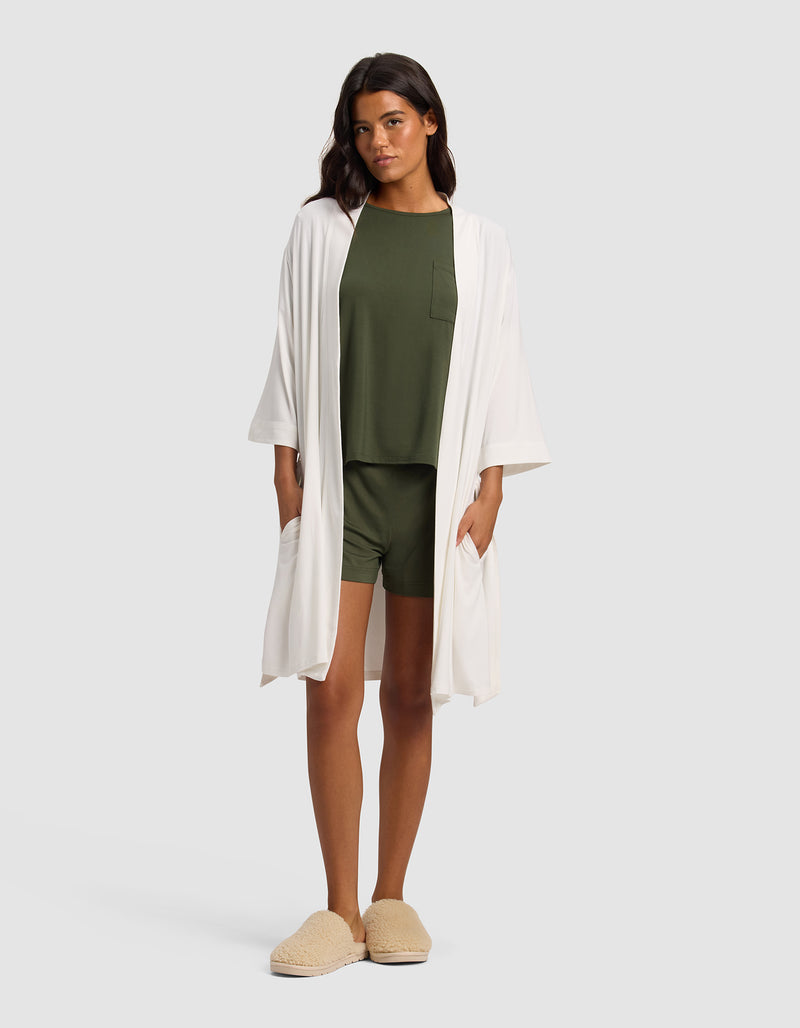 A woman with long dark hair wears the Cozy Earth Women's Bamboo Stretch-Knit Kimono Robe in white over an olive green pajama set with shorts and top, paired with beige slippers, standing against a plain light background.
