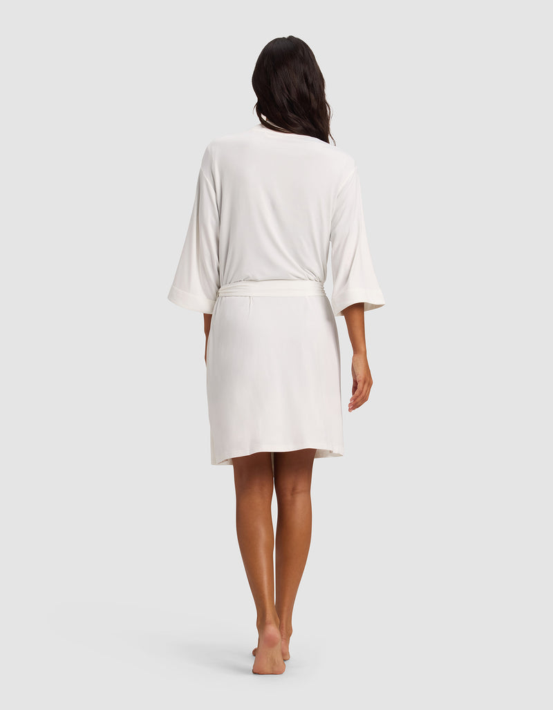 A woman with long dark hair stands barefoot, facing away, wearing the Cozy Earth Women's Bamboo Stretch-Knit Kimono Robe—a knee-length white robe with loose sleeves and a belt—against a plain light gray background.