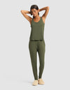 A woman stands with her arms raised, modeling the Cozy Earth Women's Bamboo Stretch-Knit Tank & Tapered Pant Set in olive green and beige slippers against a plain light background.|Color:Olive