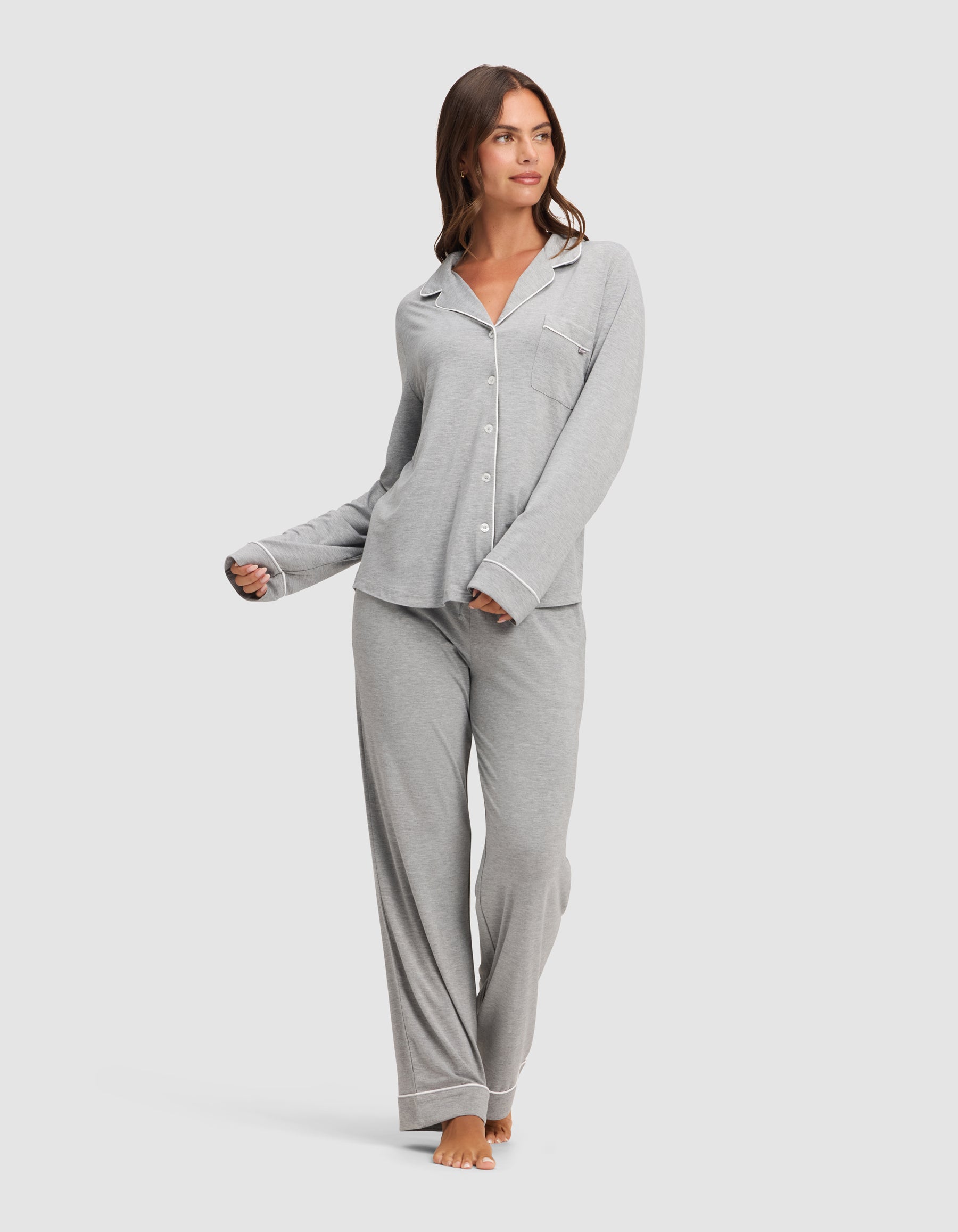 A woman stands barefoot against a plain white background, wearing the HIDE Women's Bamboo Stretch-Knit Long Sleeve Pajama Top in light gray, paired with matching loose pants. |Color:Grey