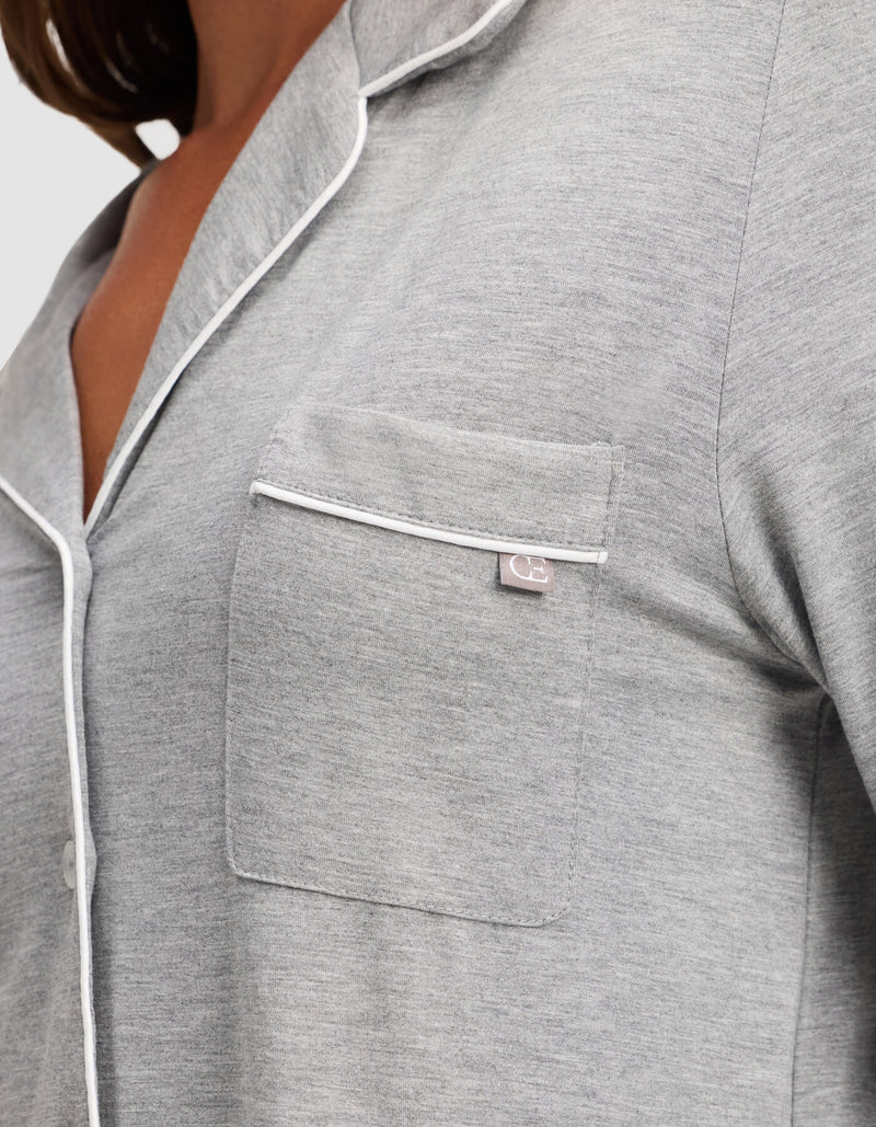 Close-up of a woman wearing Cozy Earth's Bamboo Stretch-Knit Long Sleeve Pajama Top in light grey, featuring white piping on the collar, pocket, and button line. A small fabric tag accents the chest pocket.