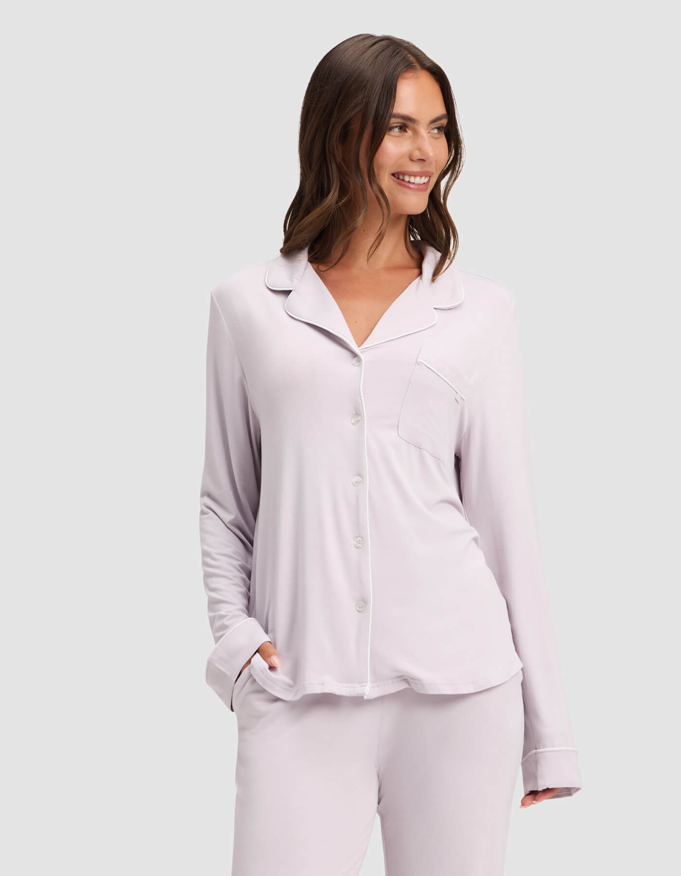 A woman smiles while wearing the Cozy Earth Women's Bamboo Stretch-Knit Long Sleeve Pajama Top in light lavender with a chest pocket, paired with matching pants. She has one hand in her pocket and stands against a plain light gray background.|Color:Lilac