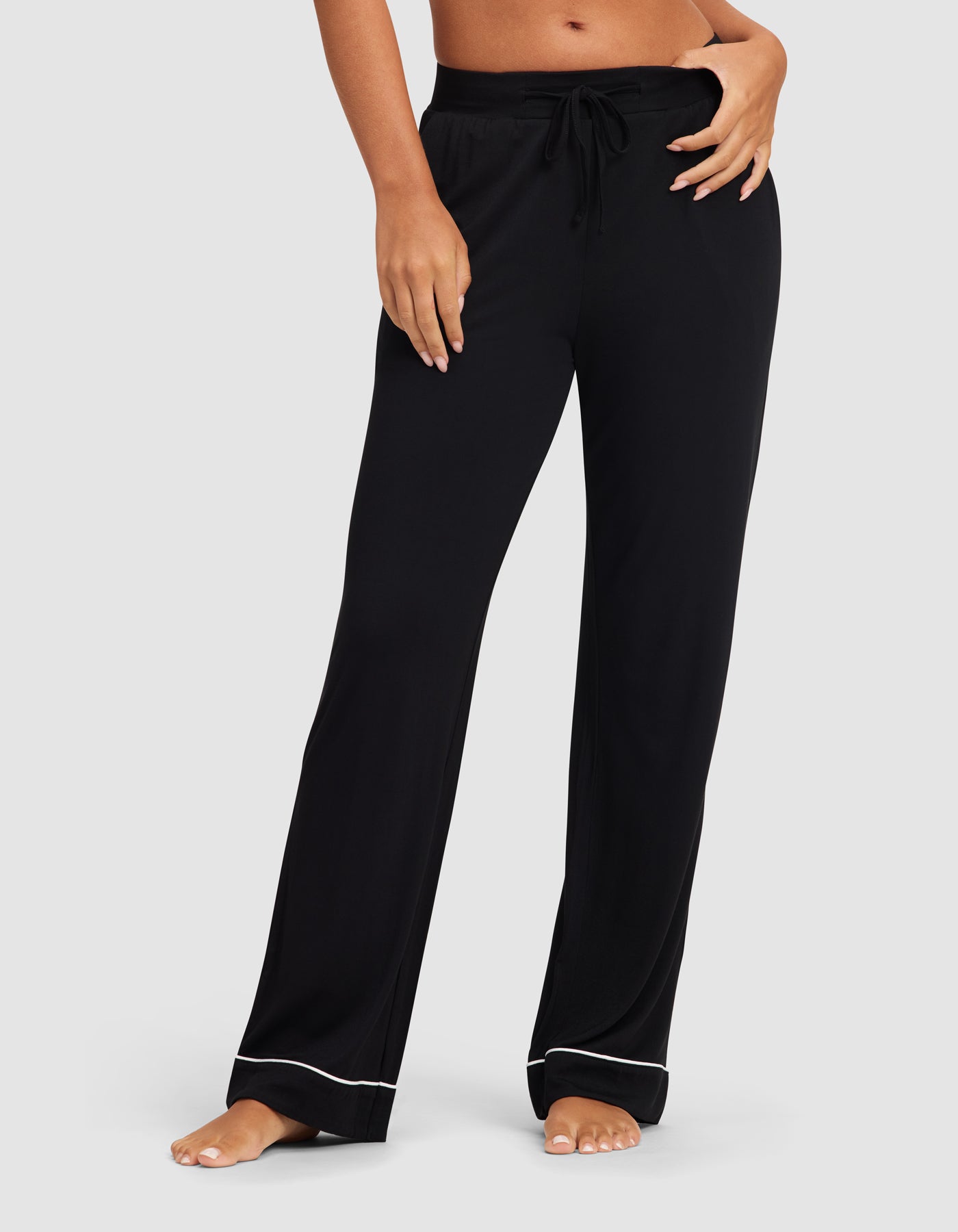 A woman models the Cozy Earth Women's Bamboo Stretch-Knit Pajama Pant in black, featuring a drawstring waist and white piping at the hem, standing barefoot against a light gray background.