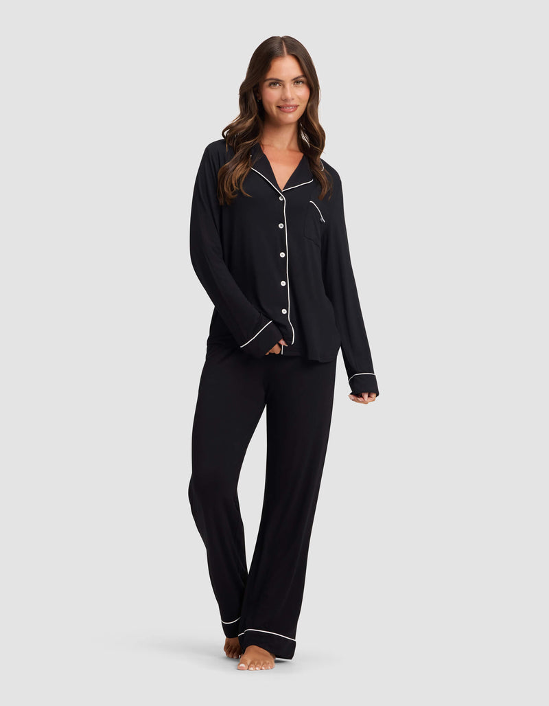 A woman with long brown hair wears Cozy Earth's Women's Bamboo Stretch-Knit Pajama Pant in black with white piping and buttons, standing barefoot and smiling against a plain light gray background.