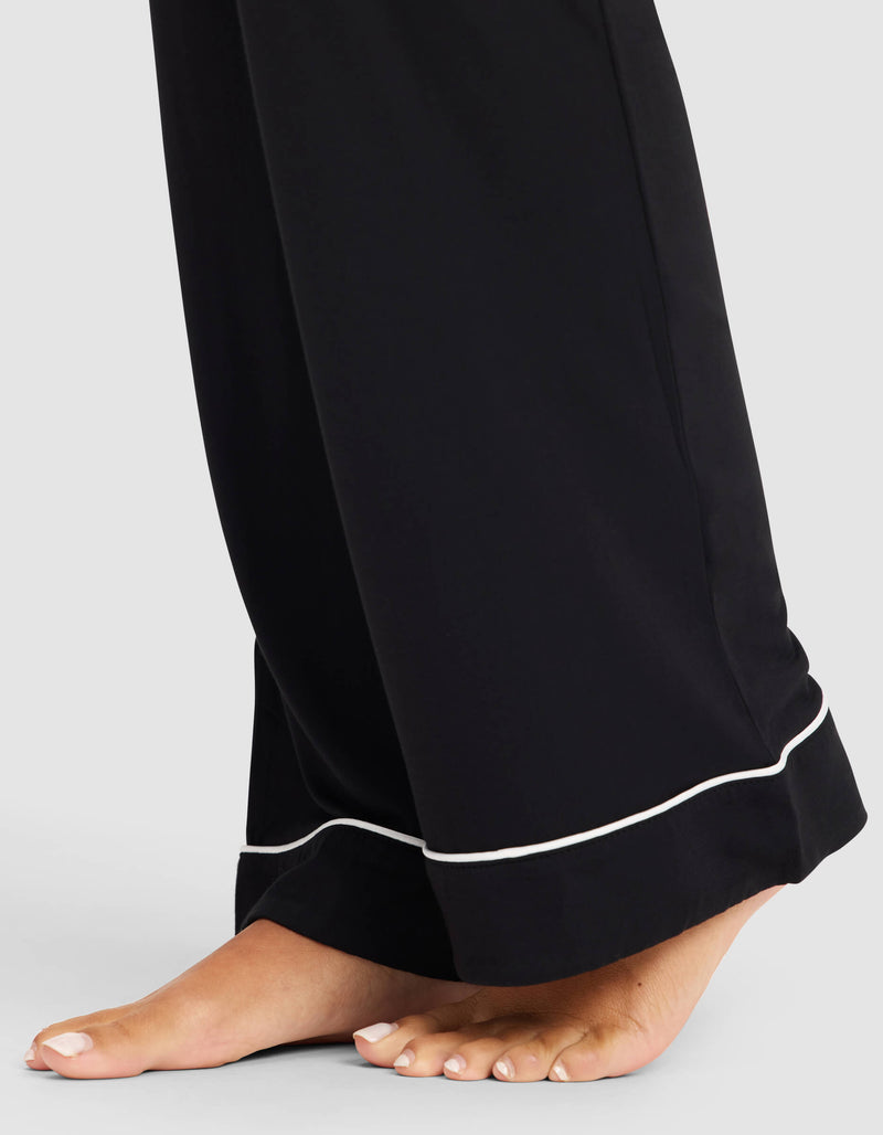 A close-up of a person’s bare feet wearing the Cozy Earth Women's Bamboo Stretch-Knit Pajama Pant in black with white piping, standing on a light gray surface.