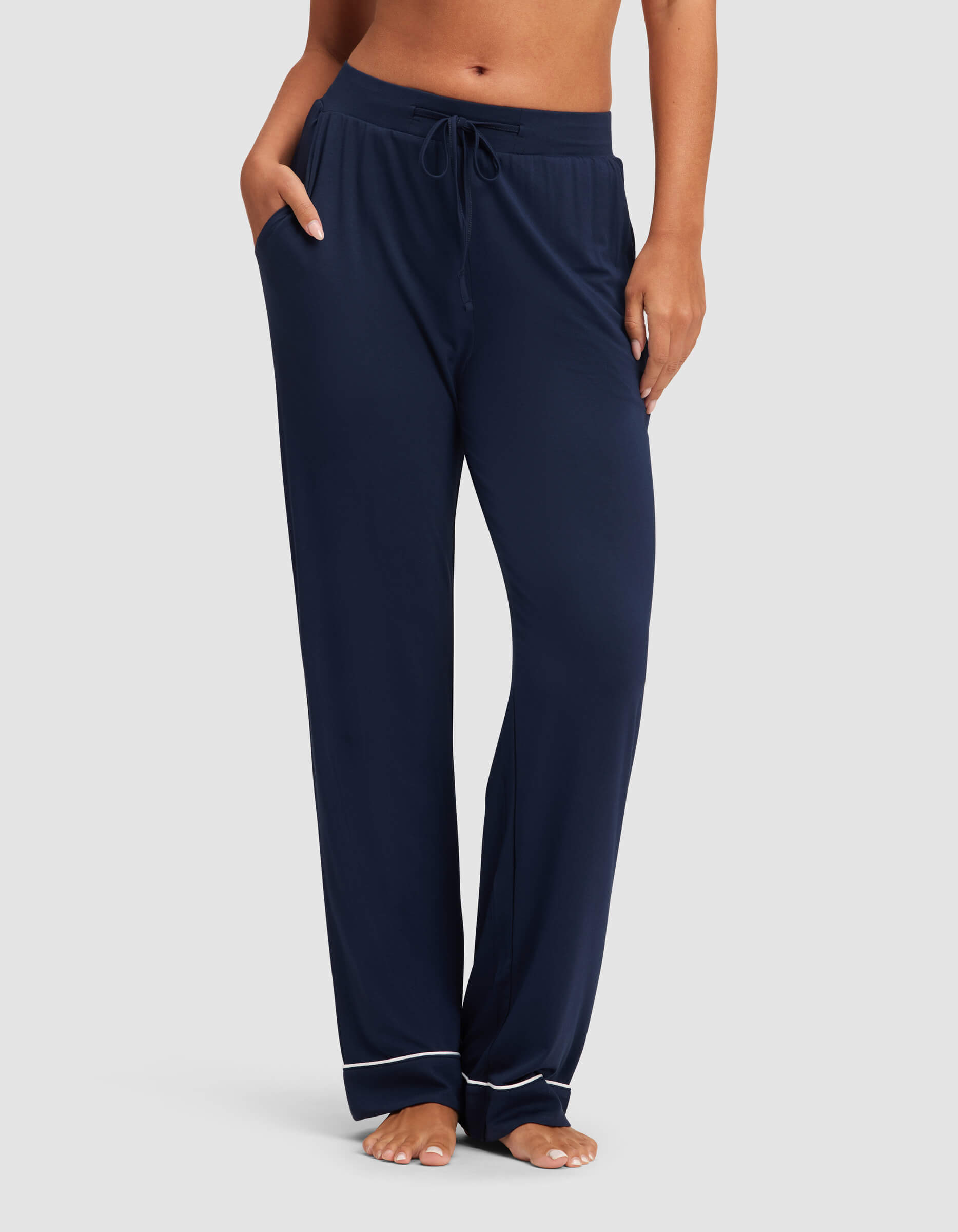 A woman stands barefoot against a plain background wearing Cozy Earth's Women's Bamboo Stretch-Knit Pajama Pant, featuring a loose fit, drawstring waist, and white piping near the hems.|Color:Navy