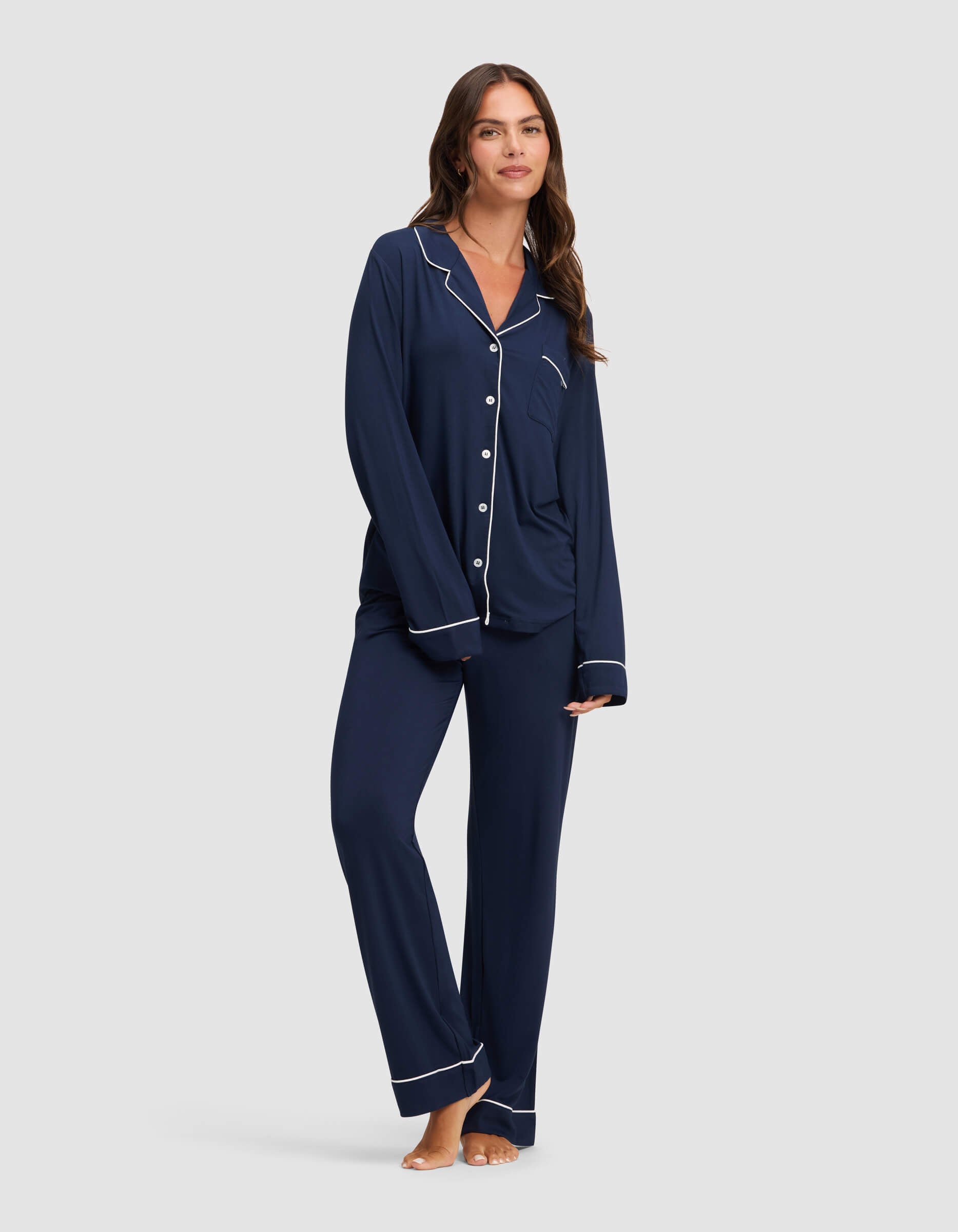 A woman with long brown hair wears Cozy Earth's Women's Bamboo Stretch-Knit Pajama Pant in navy with white piping and buttons, standing barefoot and smiling against a plain light gray background.
