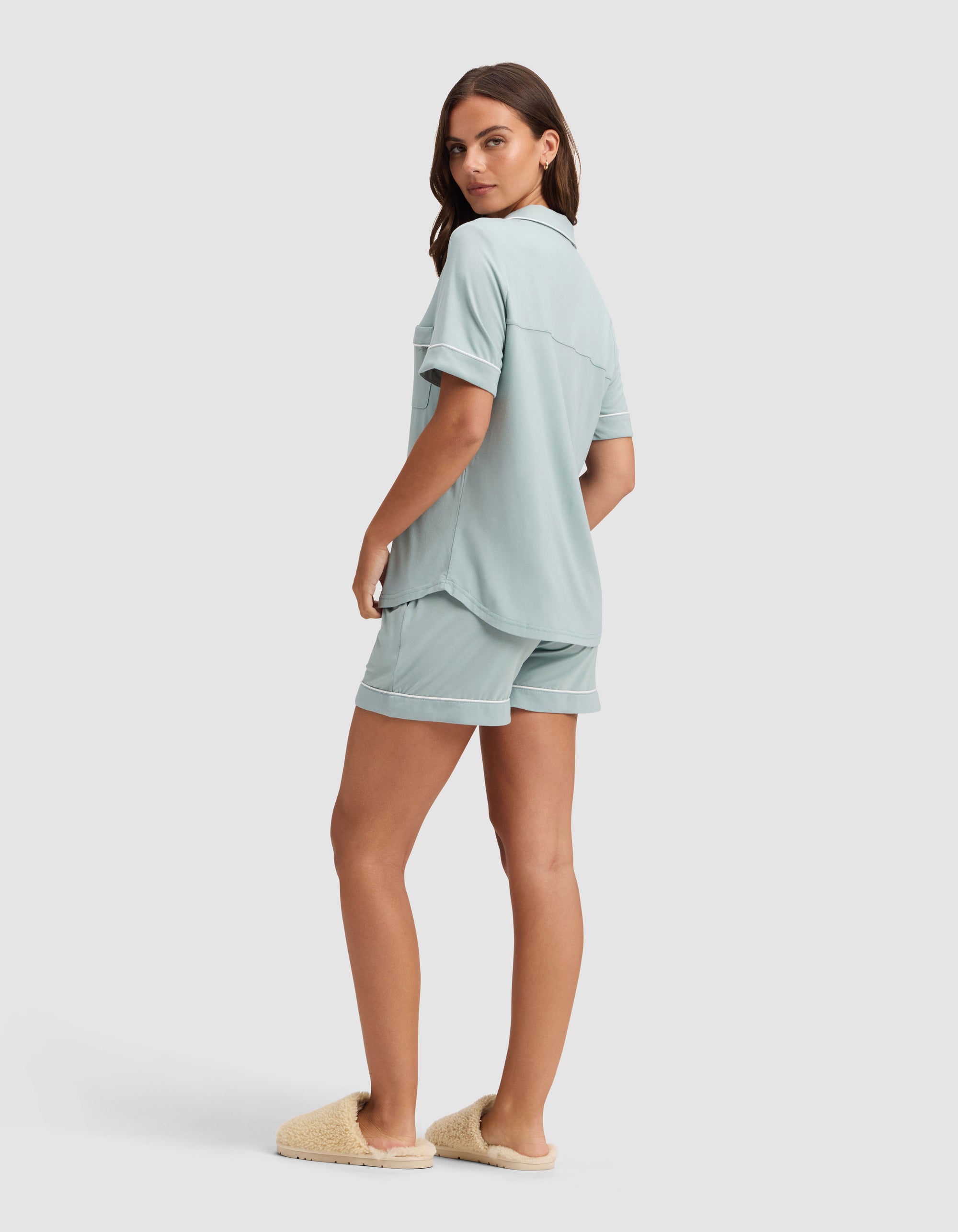 A woman, turned slightly to the side, wears the Cozy Earth Women's Bamboo Stretch-Knit Short Sleeve Pajama Set in light blue with beige slippers, posed against a plain light background. 