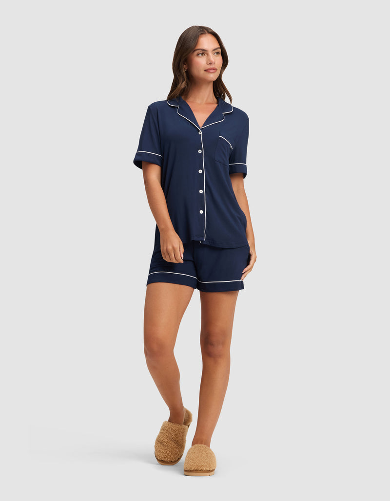 A woman wears the Cozy Earth Women's Bamboo Stretch-Knit Short Sleeve Pajama Set in navy with white piping and shorts, paired with tan fuzzy slippers, looking relaxed against a plain light background.