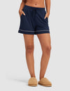 A woman wears Cozy Earth Women's Bamboo Stretch-Knit Pajama Shorts in navy with white piping, hands in pockets, and tan fuzzy slippers, standing against a plain light background.|Color:Navy