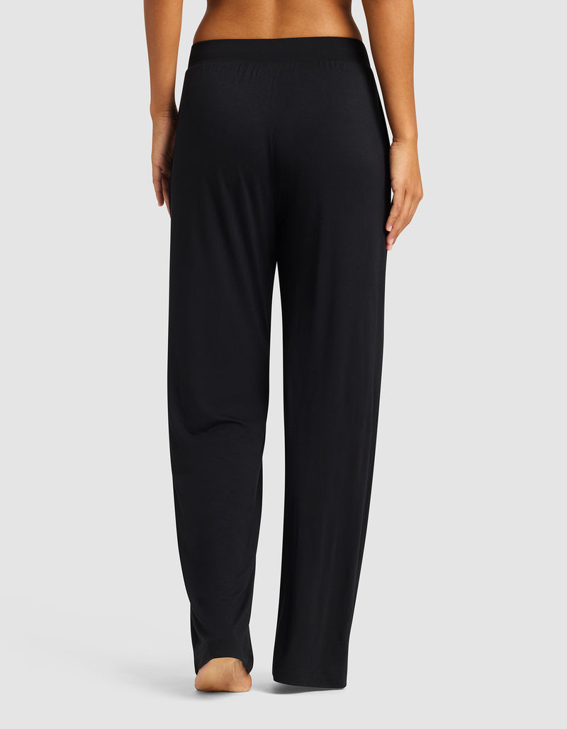 A person stands barefoot, facing away, wearing Cozy Earth’s Women's Bamboo Stretch-Knit Pant in black with an elastic waistband, set against a plain light gray background.