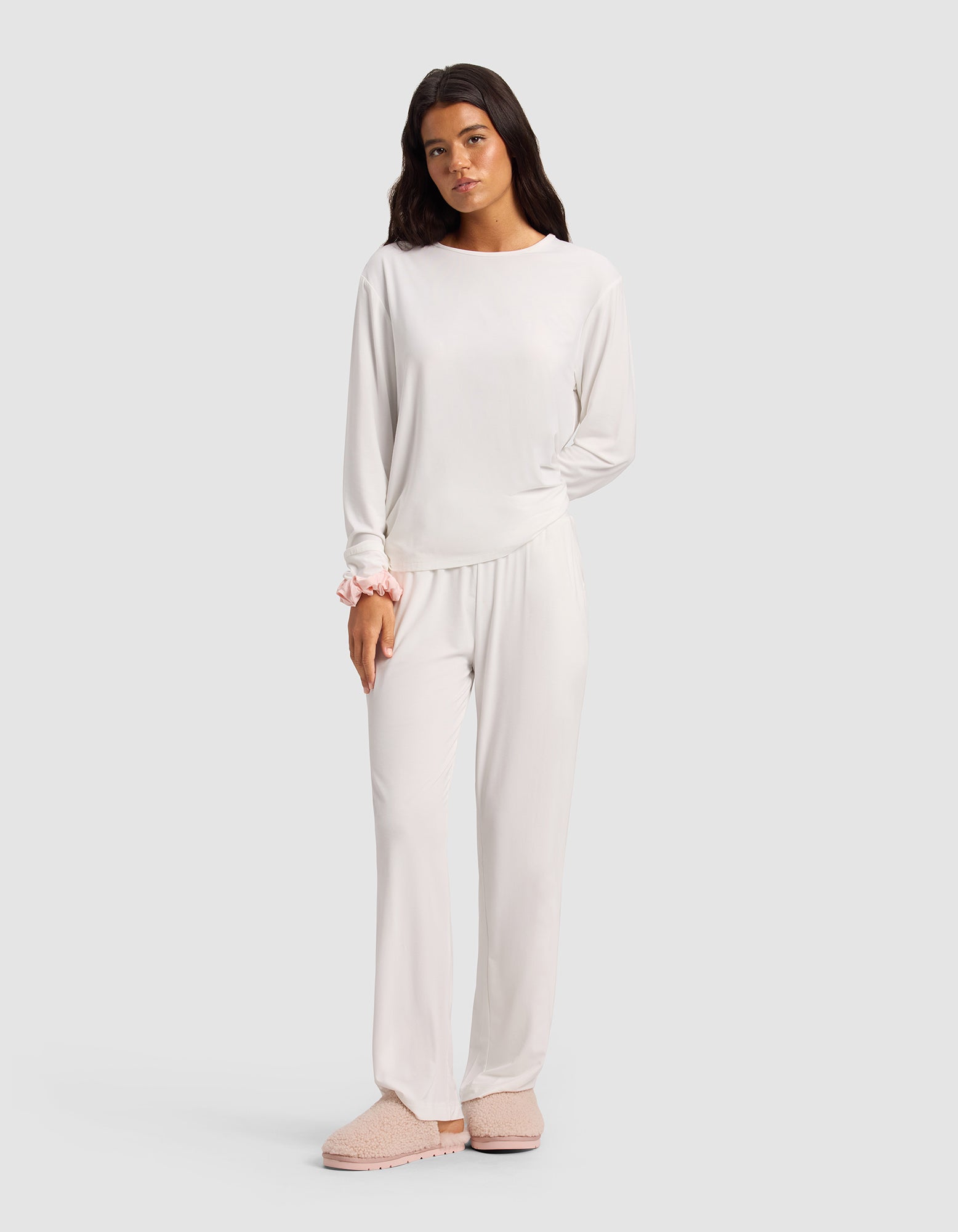 A woman stands against a plain background, wearing Cozy Earth's Women's Bamboo Stretch-Knit Pant with a matching long-sleeve top, light pink slippers, and a pink scrunchie on her wrist.