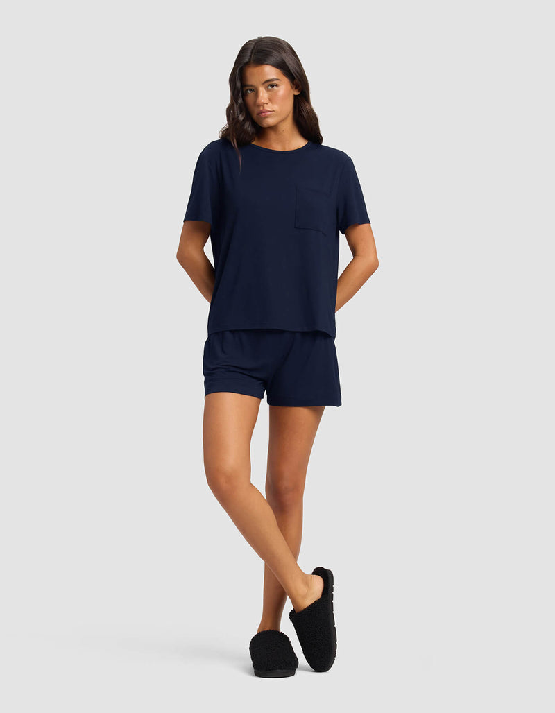 A woman poses against a plain background wearing the Cozy Earth Women's Bamboo Stretch-Knit Lounge Tee & Short Set in navy, paired with black slippers and her hands in her pockets.