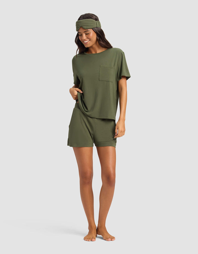 A woman stands barefoot and smiling, wearing Cozy Earth's Women's Bamboo Stretch-Knit Lounge Tee & Short Set in olive green with a matching headband, against a plain light background.