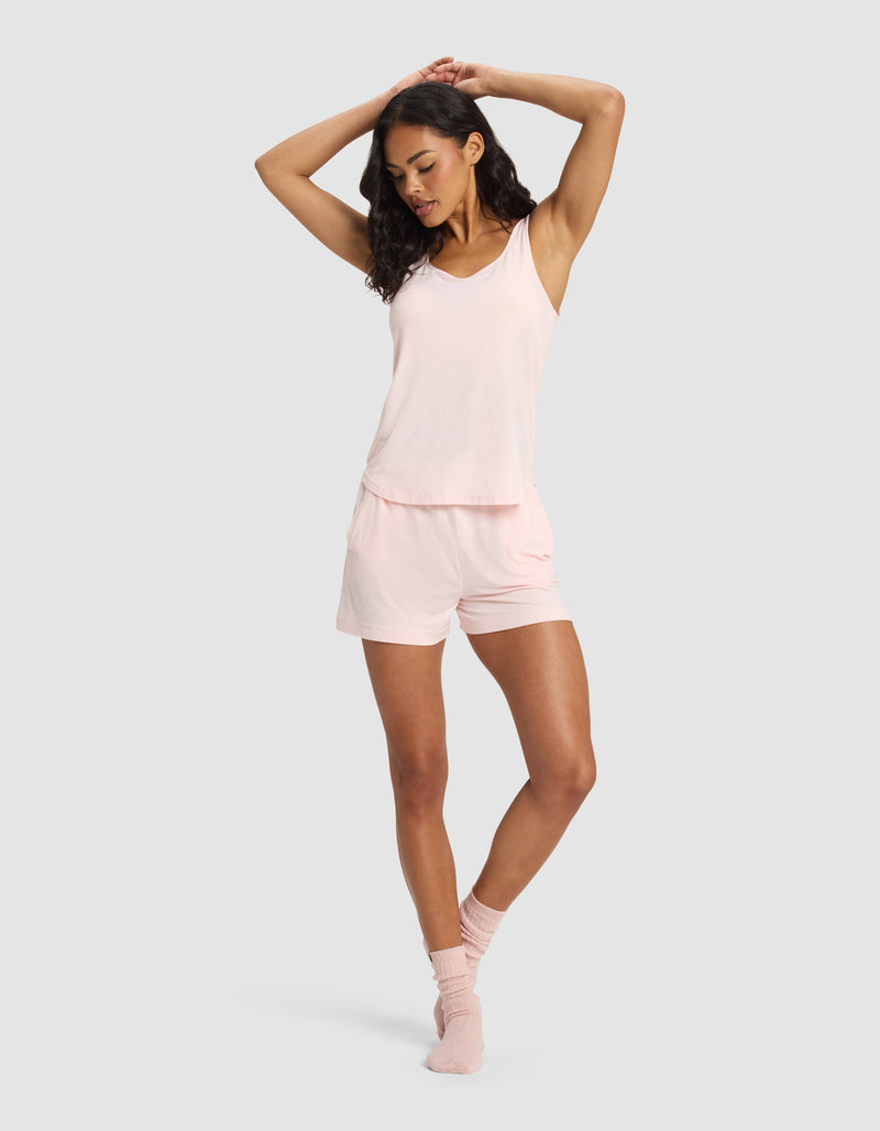 A woman stands with arms raised, wearing the Cozy Earth Women's Bamboo Stretch-Knit Lounge Tank in light pink. She pairs it with matching shorts and ankle socks, posing against a plain light gray background with a relaxed expression.