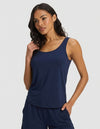 A woman wears the Cozy Earth Women's Bamboo Stretch-Knit Tank & Short Set in navy blue, standing against a light gray background with one hand in her pocket and the other touching her hair, smiling softly.|Color:Navy
