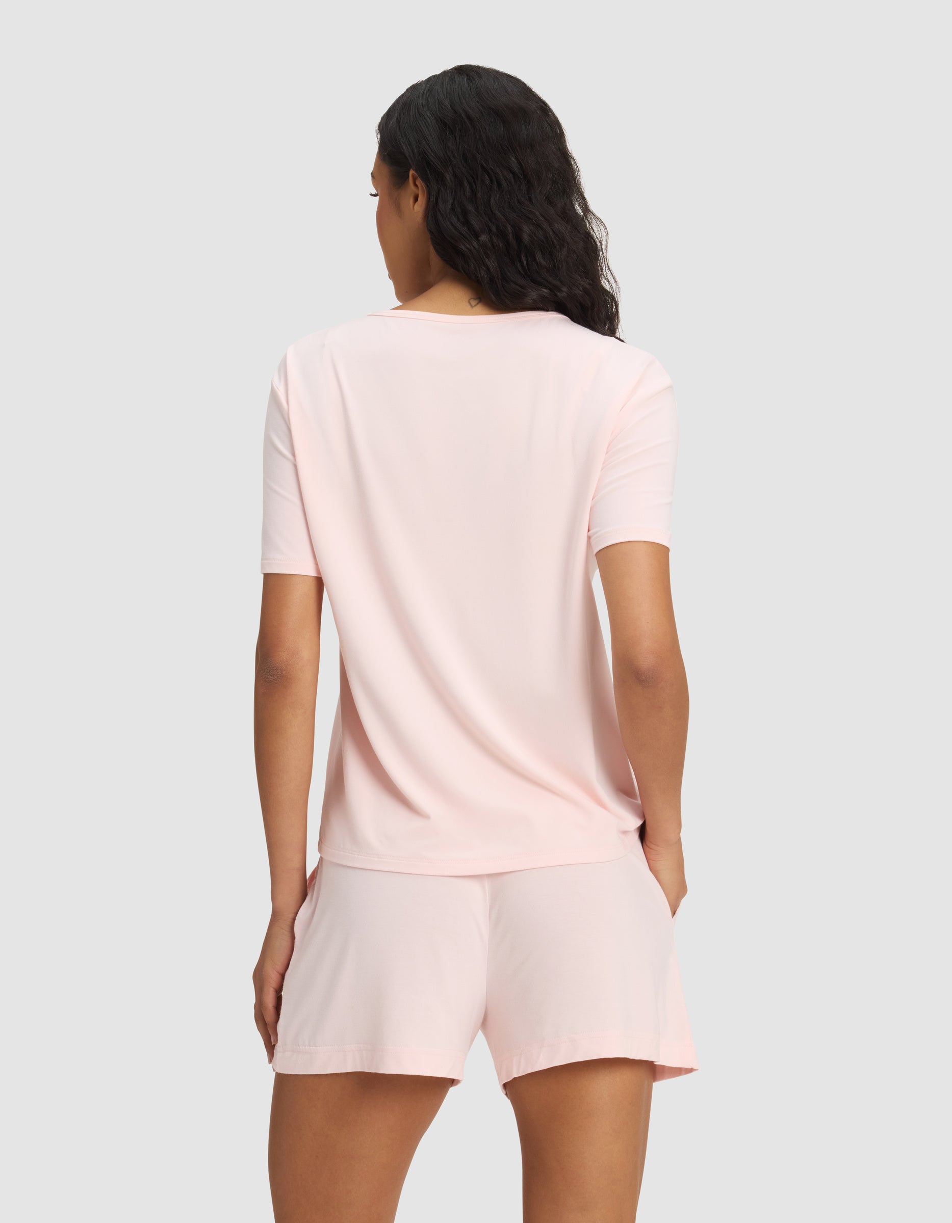 A woman with wavy dark hair is seen from the back, modeling the Cozy Earth Women's Bamboo Stretch-Knit Lounge Tee in light pink, paired with matching shorts against a plain light gray background.|Color:Blossom