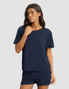 A woman with long dark hair models the Cozy Earth Women's Bamboo Stretch-Knit Lounge Tee & Short Set in navy, featuring a short-sleeve top with chest pocket and matching shorts, against a plain light gray background.|Color:Navy
