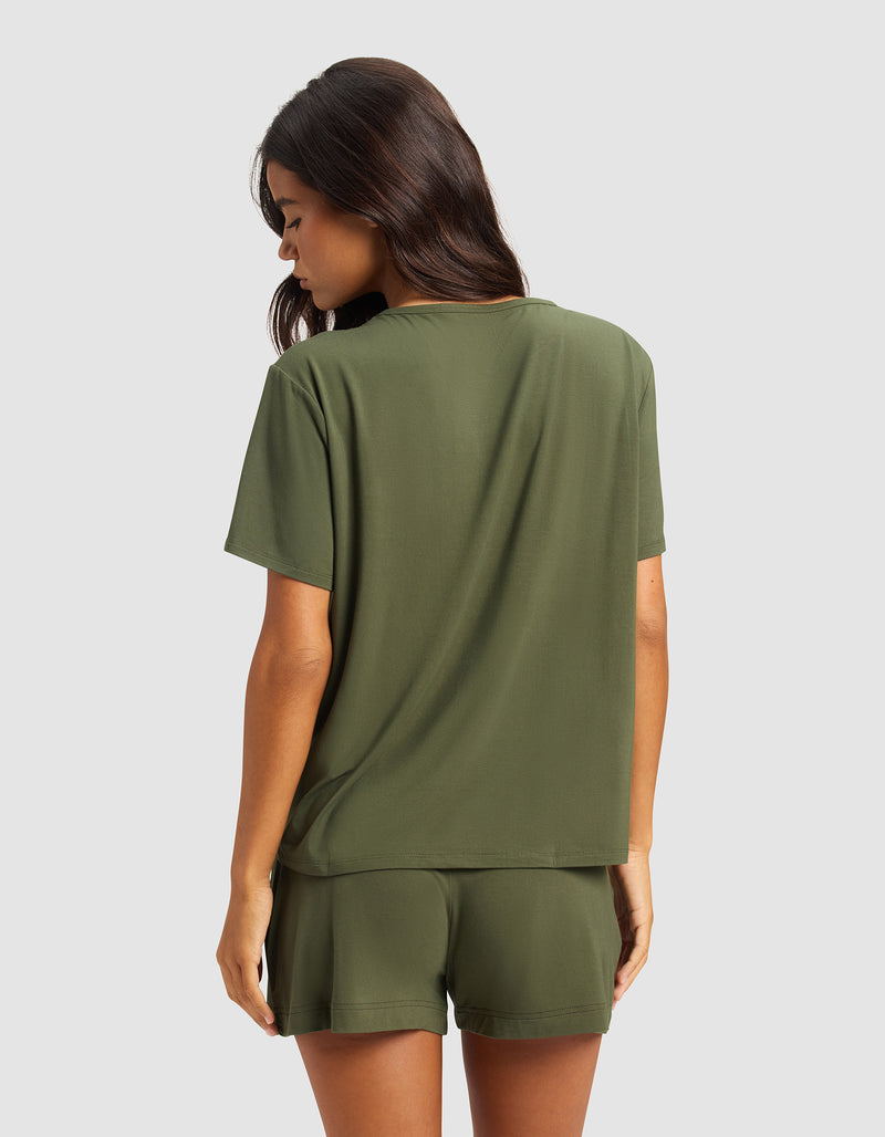 A woman with long brown hair faces away, wearing Cozy Earth's Women's Bamboo Stretch-Knit Lounge Tee in olive green, paired with matching shorts, set against a plain light gray background.