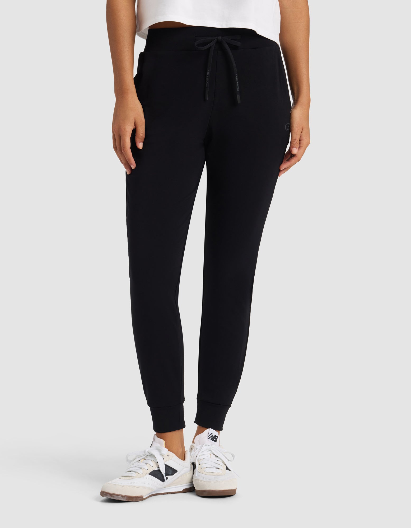 A person stands against a light gray background with hands in pockets, wearing Cozy Earth's Women's Brushed Bamboo Jogger Pant in black, paired with a white shirt and white sneakers.