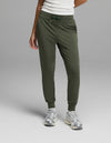 A person is shown from the waist down, wearing Cozy Earth’s Women’s Brushed Bamboo Jogger in olive green with a white top and silver sneakers, standing against a plain gray background. Only the lower body and part of one arm are visible.|Color:Olive