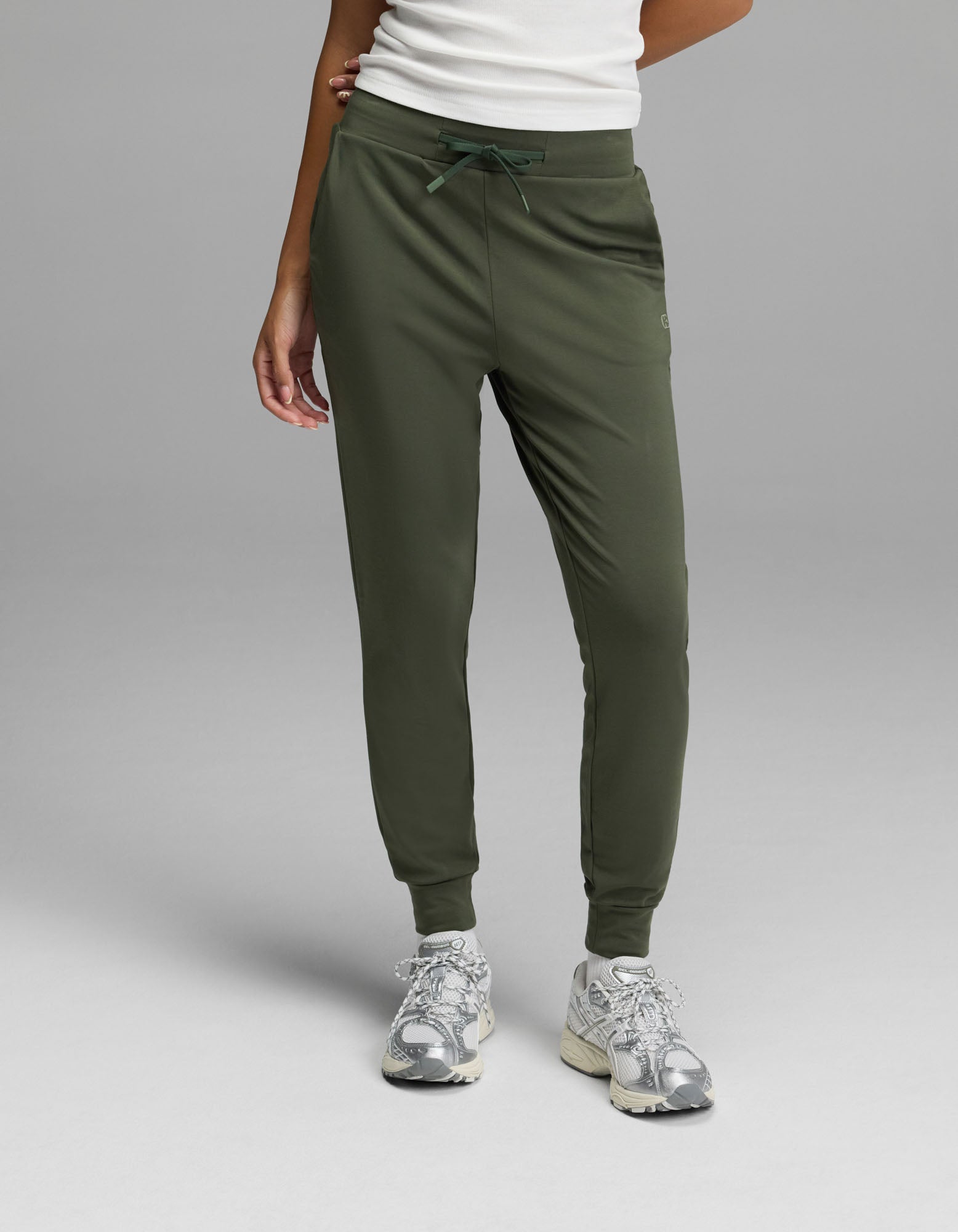 A person is shown from the waist down, wearing Cozy Earth’s Women’s Brushed Bamboo Jogger in olive green with a white top and silver sneakers, standing against a plain gray background. Only the lower body and part of one arm are visible.|Color:Olive
