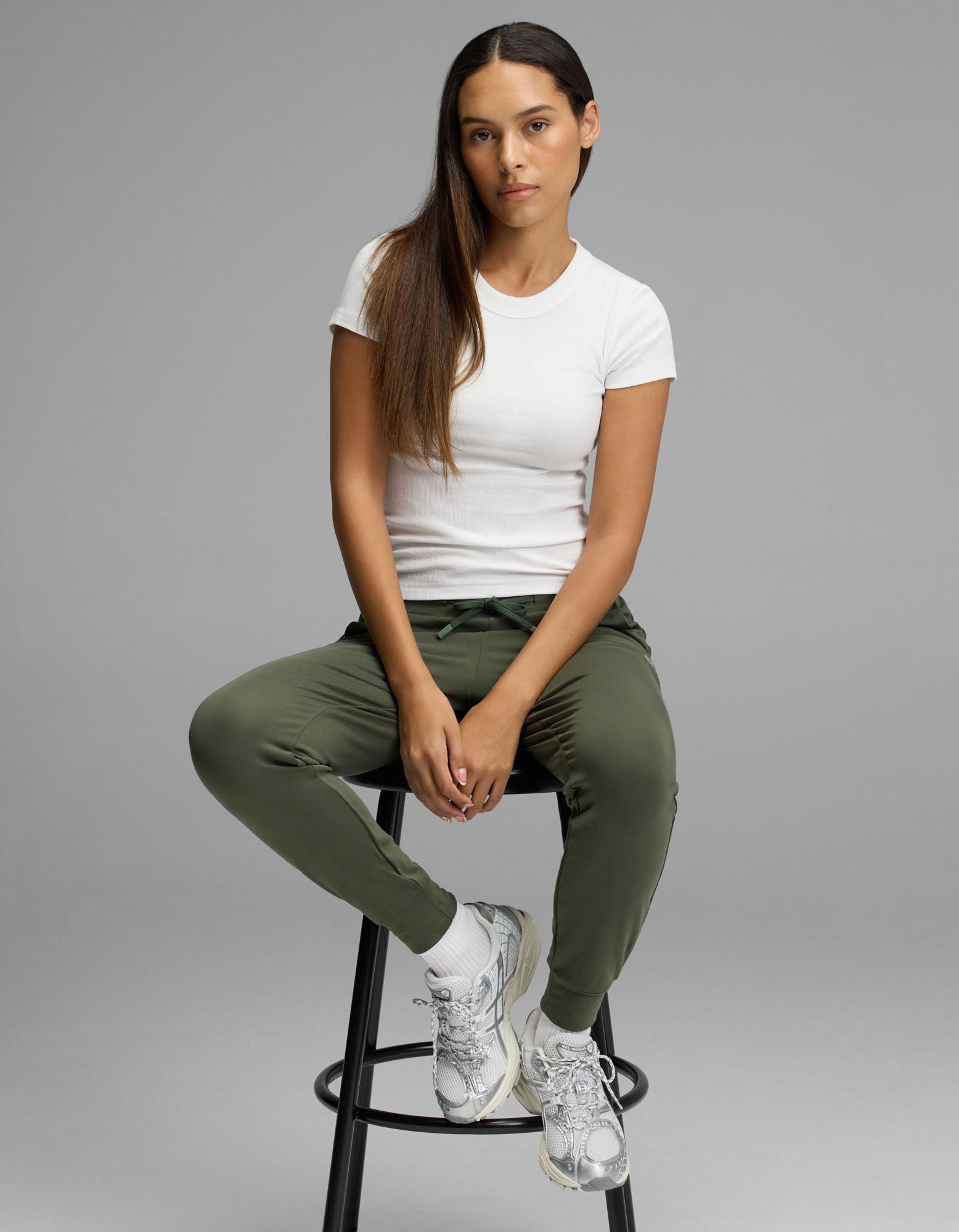 A woman with long brown hair sits on a black stool, wearing the Cozy Earth Women’s Brushed Bamboo Jogger in olive green, paired with a white t-shirt, white socks, and silver sneakers. She looks at the camera with a neutral expression against a plain gray background.
